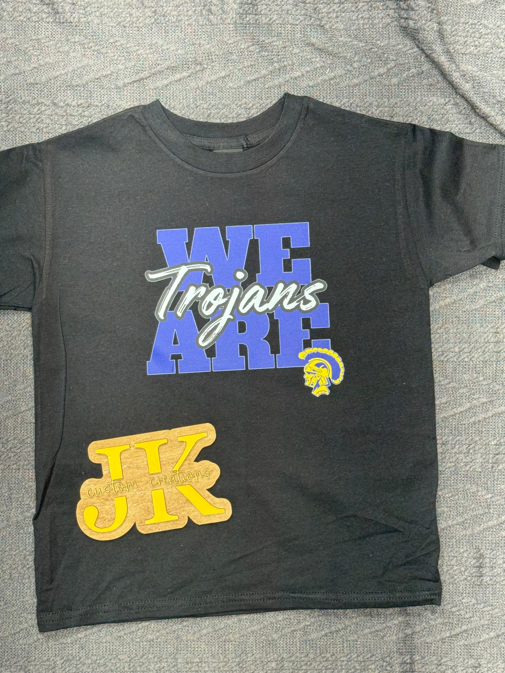 Black t-shirt with 'WE Trojans ARE' text and JK logo on a gray surface
